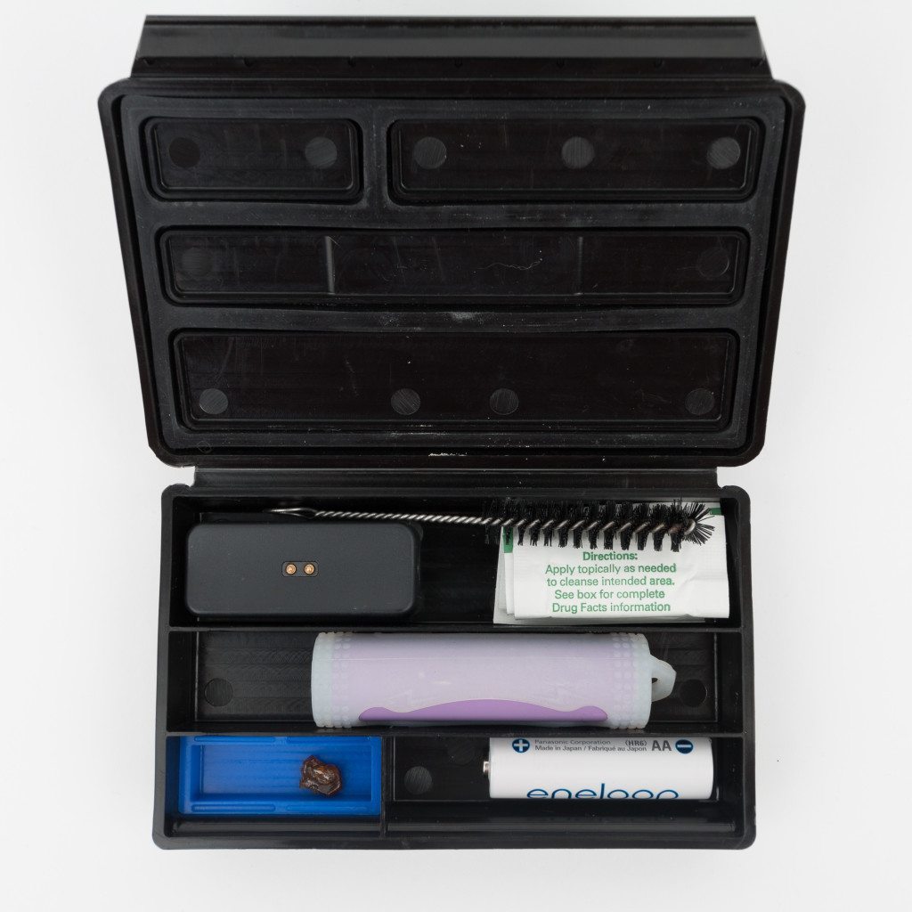 Top down view of the Bhanga Box open with Pax 2 charging block, alcohol wipes, cleaning brush, an 18650 battery, one AA battery, and a silicone insert in the smallest compartment